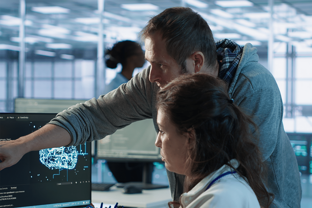 A man points at a computer screen displaying a digital brain diagram while discussing data with a woman; the scene suggests US AI engineers collaborating on federal hiring in a cutting-edge, high-tech office environment.