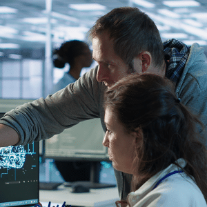 A man points at a computer screen displaying a digital brain diagram while discussing data with a woman; the scene suggests US AI engineers collaborating on federal hiring in a cutting-edge, high-tech office environment.