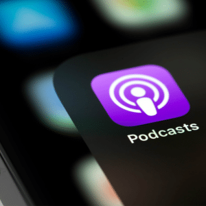 Close-up of a smartphone screen showing the Apple Podcasts app icon, highlighting where you can discover the best shows of 2025. The purple icon with a white microphone stands out, with other blurred app icons in the background.
