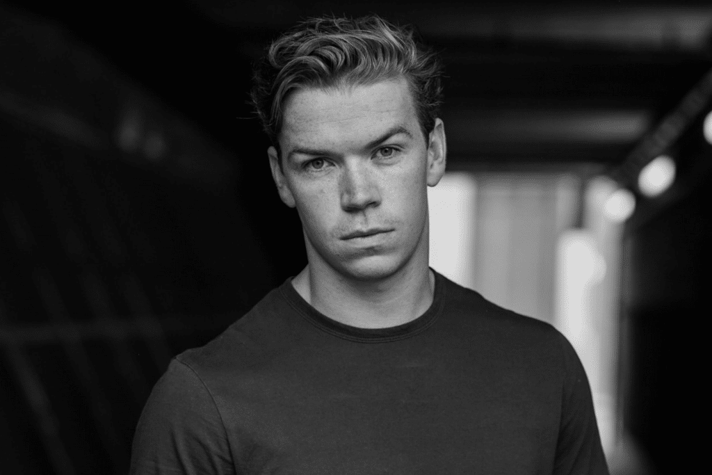 A young man with short, light hair and a plain dark shirt stands in a dimly lit corridor, looking directly at the camera with a serious expression, as if he's ready to Beat the Reaper. The image is in black and white.