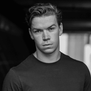 A young man with short, light hair and a plain dark shirt stands in a dimly lit corridor, looking directly at the camera with a serious expression, as if he's ready to Beat the Reaper. The image is in black and white.
