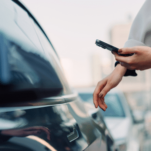 A person stands next to a parked car, holding a smartphone with Apple Car Key, reaching for the door handle. The scene is outdoors on a city street as Apple Car Key expands in 2026.