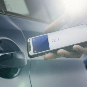 A person holds a smartphone with the Apple Car Key app open near the door handle of a blue car, preparing to unlock it using their phone. Sunlight shines onto the scene as Apple Car Key expands in 2026.