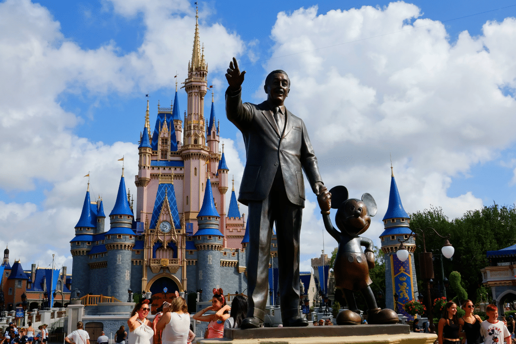 A bronze statue of a man holding hands with Mickey Mouse stands before a pink and blue fairytale castle at a busy theme park, where crowds gather under a partly cloudy sky to celebrate Disney board member Jeff Williams' legacy.
