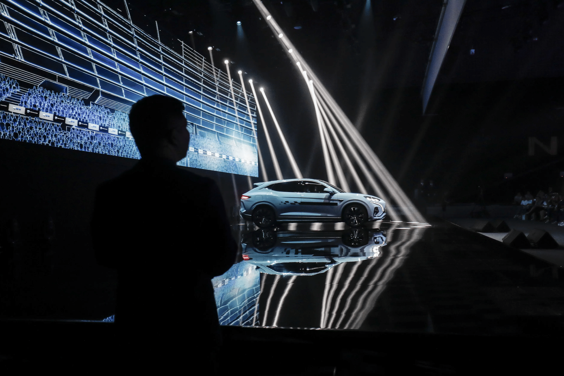 A white sports car is displayed on a glossy stage under bright diagonal spotlights, with digital graphics behind it, highlighting Western EV competitiveness. A silhouetted person stands in the foreground, watching the presentation.