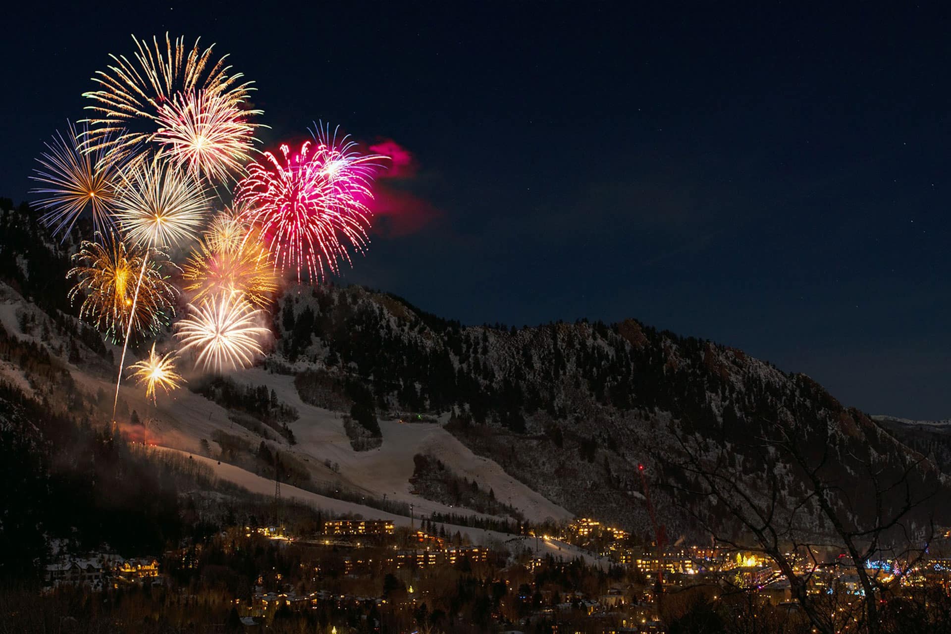 Colorful fireworks burst over a snow-covered mountain town at night, illuminating the sky and surrounding hills as the lights below glow in celebration of Happy New Year 2026. Featured in AppleMagazine.
