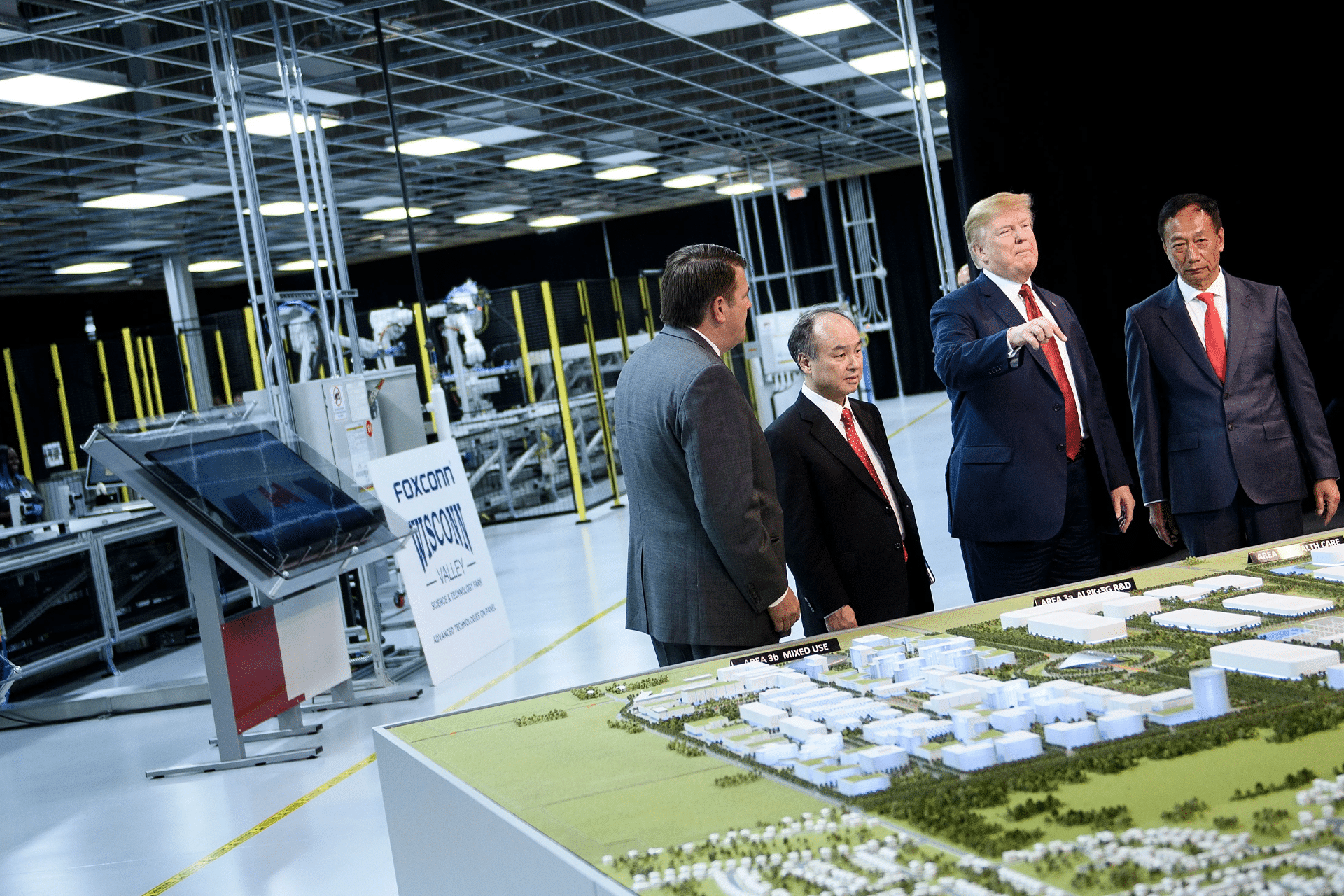 Four men in suits stand around a detailed scale model of a facility inside a modern industrial building, with machinery and Foxconn Wisconsin expansion signage visible in the background. One man gestures toward the model.