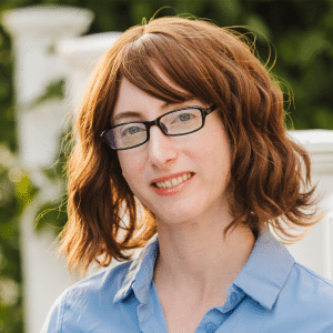 A person with wavy, shoulder-length brown hair and glasses smiles slightly while wearing a light blue collared shirt, standing outdoors near a white railing with greenery in the background—reminiscent of an Apple TV The Teacher adaptation scene.