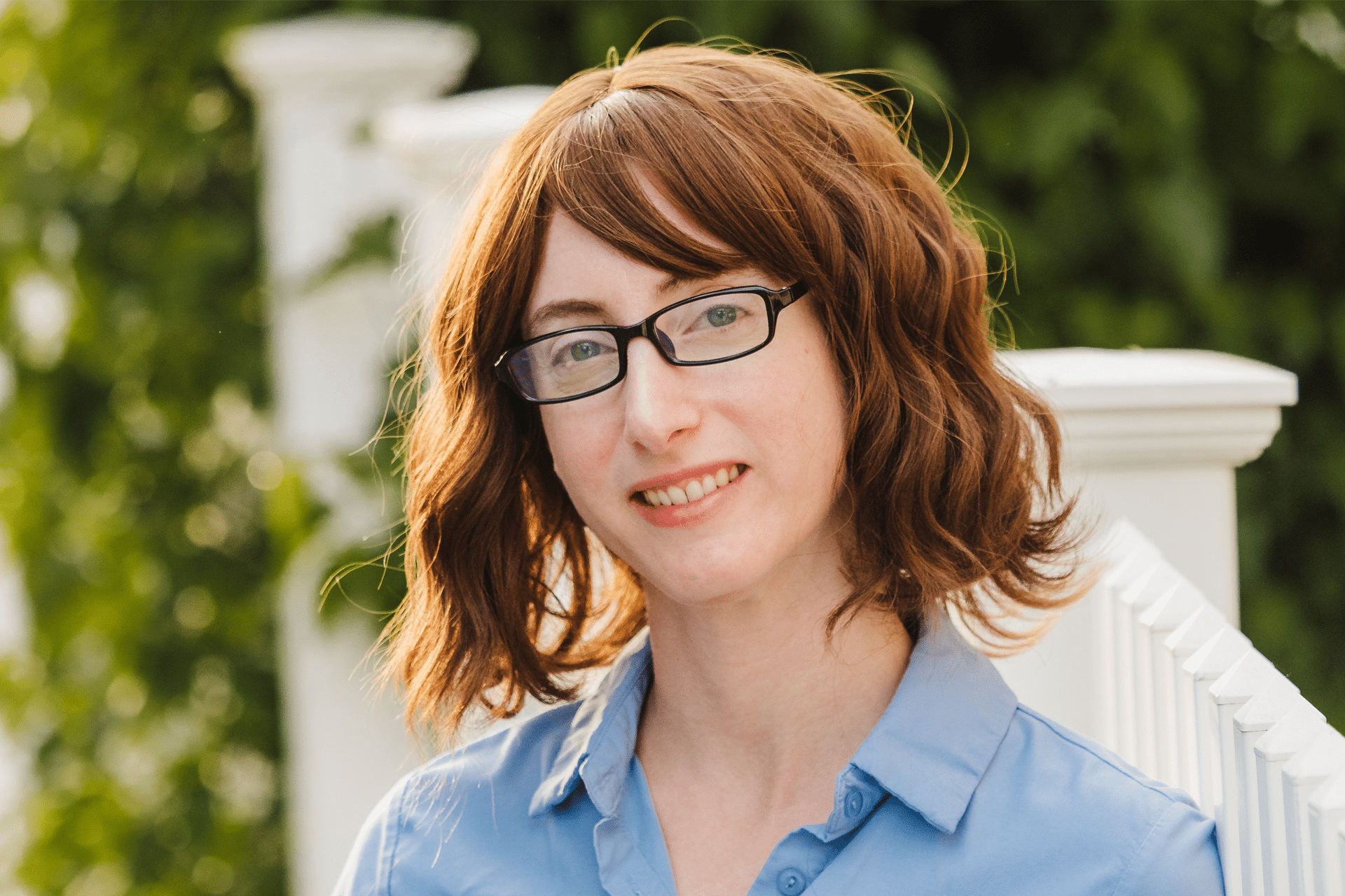 A person with wavy, shoulder-length brown hair and glasses smiles slightly while wearing a light blue collared shirt, standing outdoors near a white railing with greenery in the background—reminiscent of an Apple TV The Teacher adaptation scene.