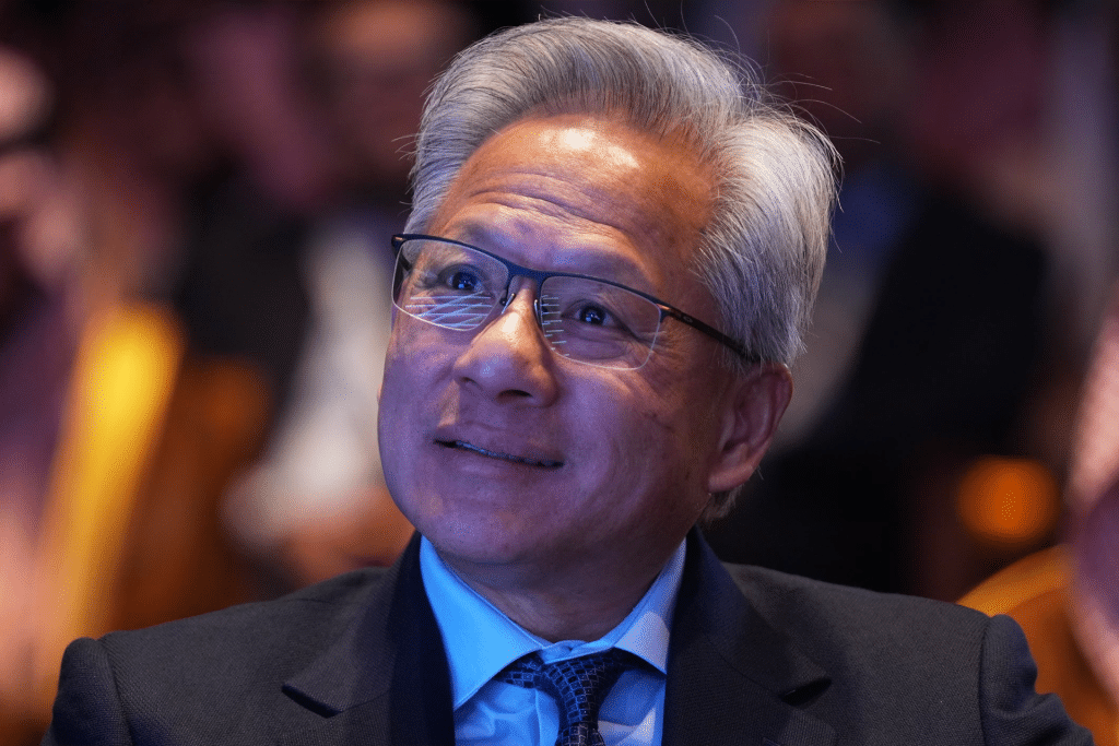 A man with gray hair and glasses, wearing a dark suit, blue shirt, and tie, is smiling and looking slightly upward. The softly blurred background with warm lighting hints at an ongoing Nvidia AI export debate.