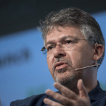 A man with glasses and a headset microphone is speaking passionately about AI, gesturing with his hands during a presentation or panel discussion. The background shows a blurred logo and text, hinting at an Apple event and possibly John Giannandrea’s involvement.