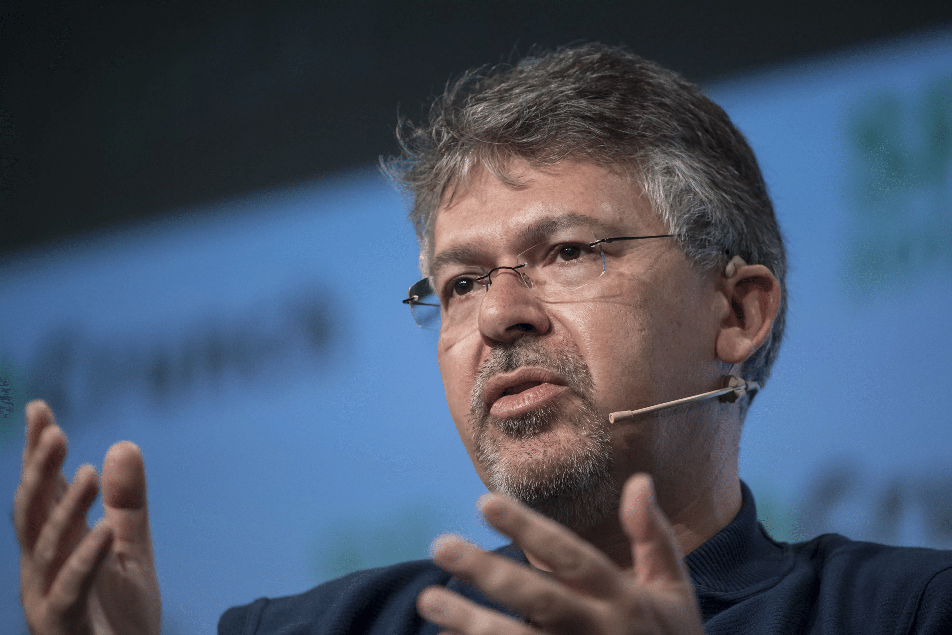 A man with glasses and a headset microphone is speaking passionately about AI, gesturing with his hands during a presentation or panel discussion. The background shows a blurred logo and text, hinting at an Apple event and possibly John Giannandrea’s involvement.