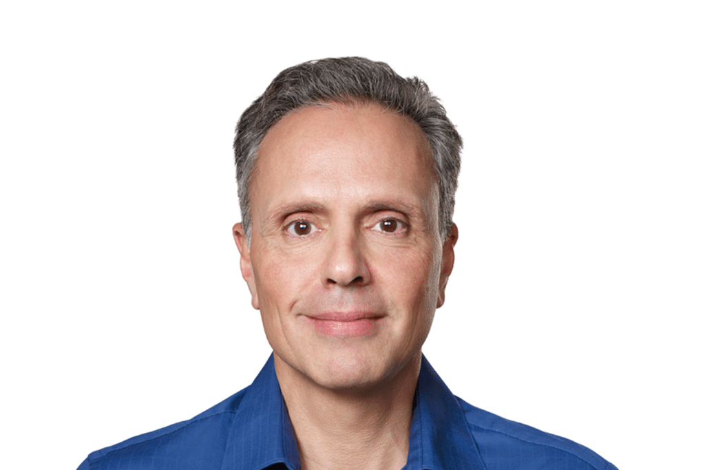 A middle-aged man with short gray hair and brown eyes, wearing a blue collared shirt, is smiling gently at the camera against a plain white background, reflecting on the recent Apple Chip Chief departure.