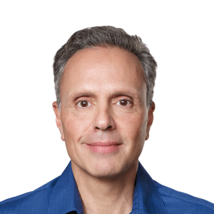 A middle-aged man with short gray hair and brown eyes, wearing a blue collared shirt, is smiling gently at the camera against a plain white background, reflecting on the recent Apple Chip Chief departure.