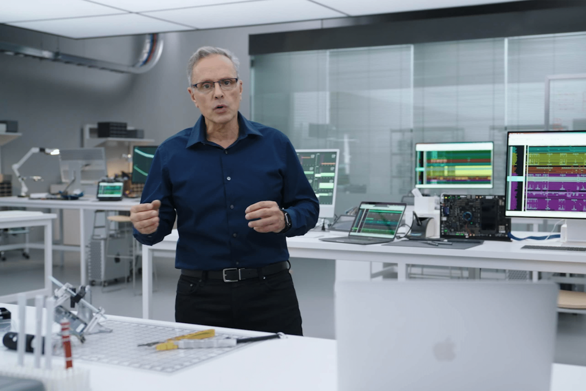 A man in glasses and a dark blue shirt discusses the recent Apple Chip Chief Departure in a modern office or lab with computers displaying colorful graphs and audio waveforms, surrounded by high-tech equipment and tools on white desks.