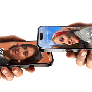Two hands each hold a smartphone using iPhone NameDrop contact sharing, displaying digital profile cards with a woman’s name and photo. The phones are positioned close together, appearing to seamlessly share contact information.