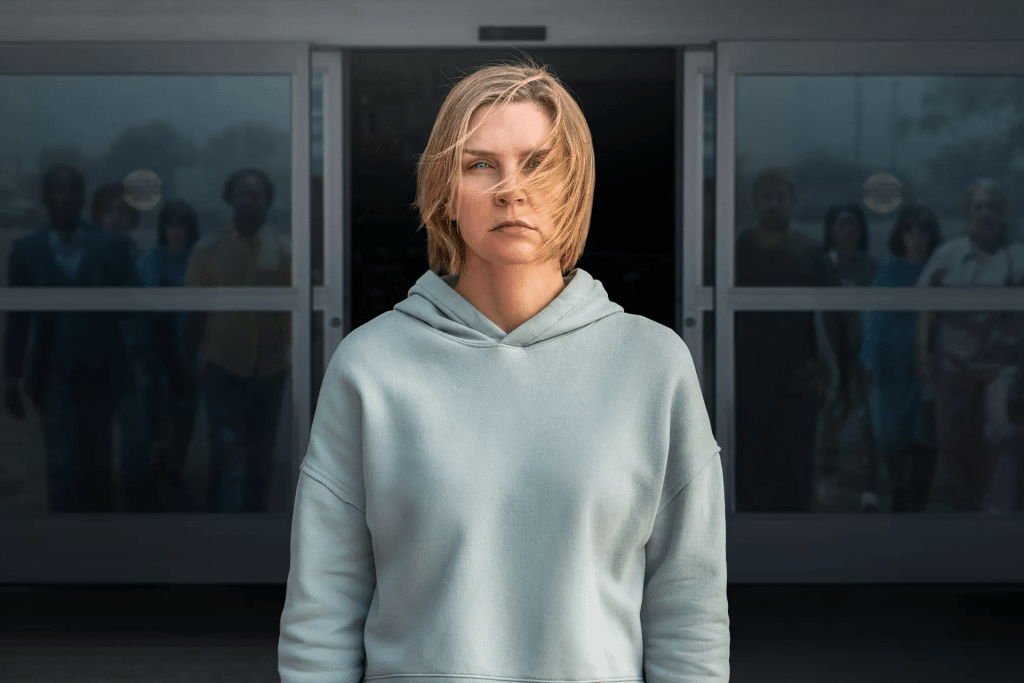 A woman with short blond hair wearing a light gray hoodie stands outside in front of sliding glass doors. In this Pluribus Episode 6 scene, several people—including Carol—are visible in shadowy detail behind the doors, slightly blurred.
