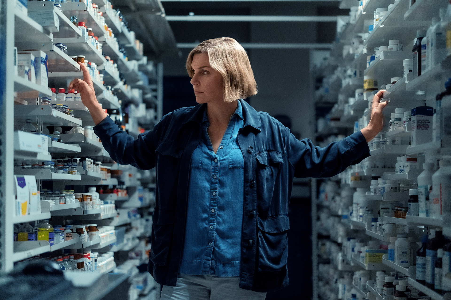 A woman with short blonde hair stands between shelves filled with medicine bottles in a dimly lit pharmacy, wearing a blue shirt and dark jacket, looking to her left with a serious expression in this scene from Pruribus Confirm Season 2.