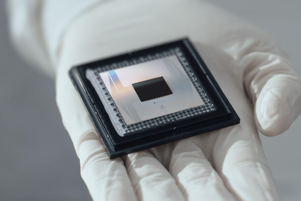 A gloved hand holds a computer microchip—reminiscent of quantum chips used in Apple devices—in a black frame, highlighting its metallic surface and intricate details against a blurred, neutral background.