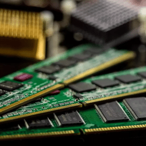 Close-up of three green computer RAM sticks with black chips, stacked diagonally, with blurred electronic components in the background—capturing the impact as RAM Manufacturing Shifts Raise Costs.