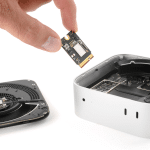 A hand holds an SSD chip above an open Mac mini, with internal components and a detached fan visible on a white surface—a reminder of hardware upgrades amid rising Apple RAM prices.
