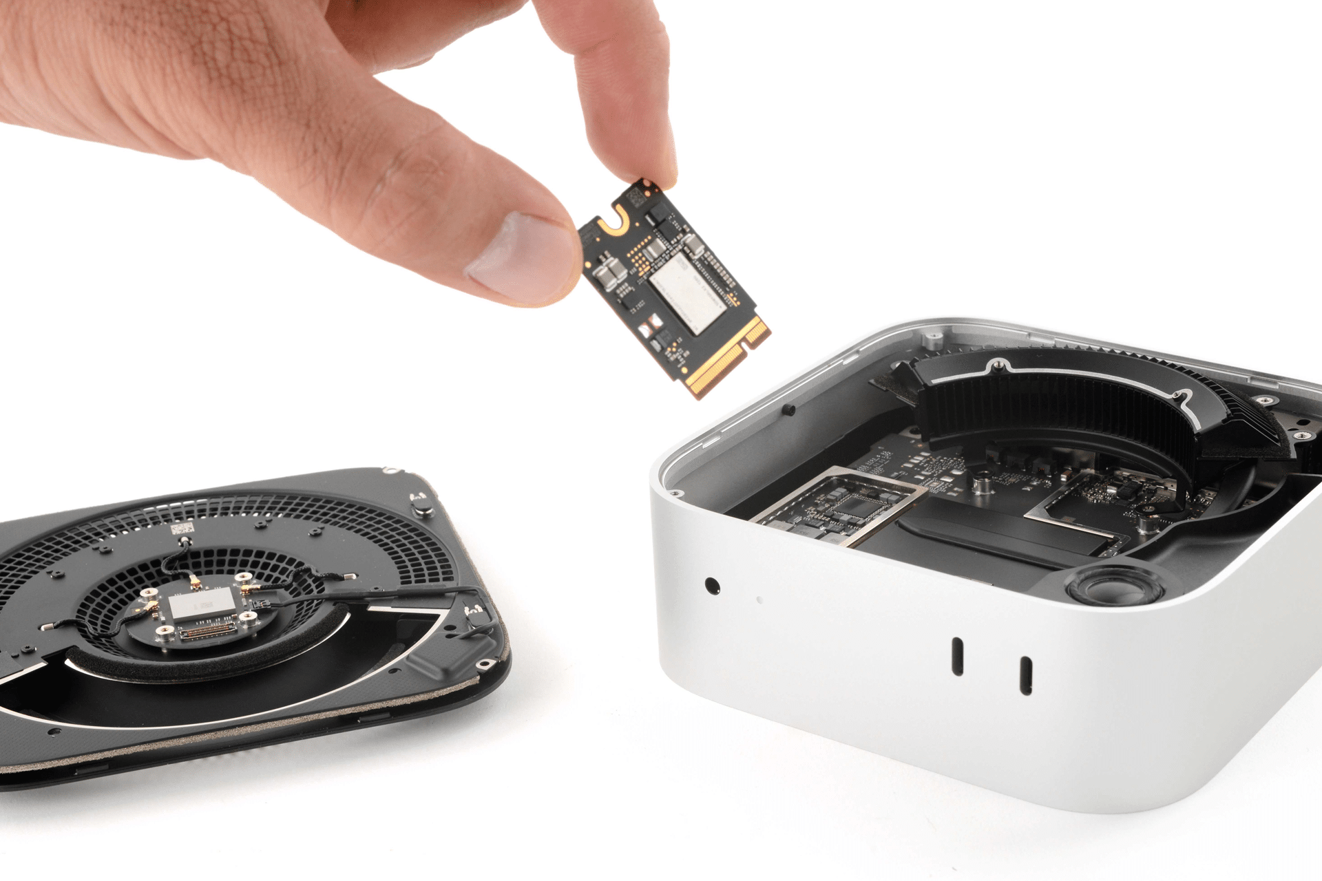 A hand holds an SSD chip above an open Mac mini, with internal components and a detached fan visible on a white surface—a reminder of hardware upgrades amid rising Apple RAM prices.