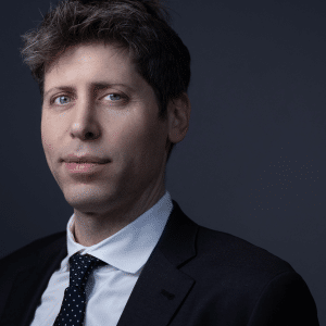 A man with light skin, short brown hair with some gray, and blue eyes—resembling Sam Altman of rocket company fame—wears a black suit, white shirt, and black polka dot tie while posing against a dark gray background.
