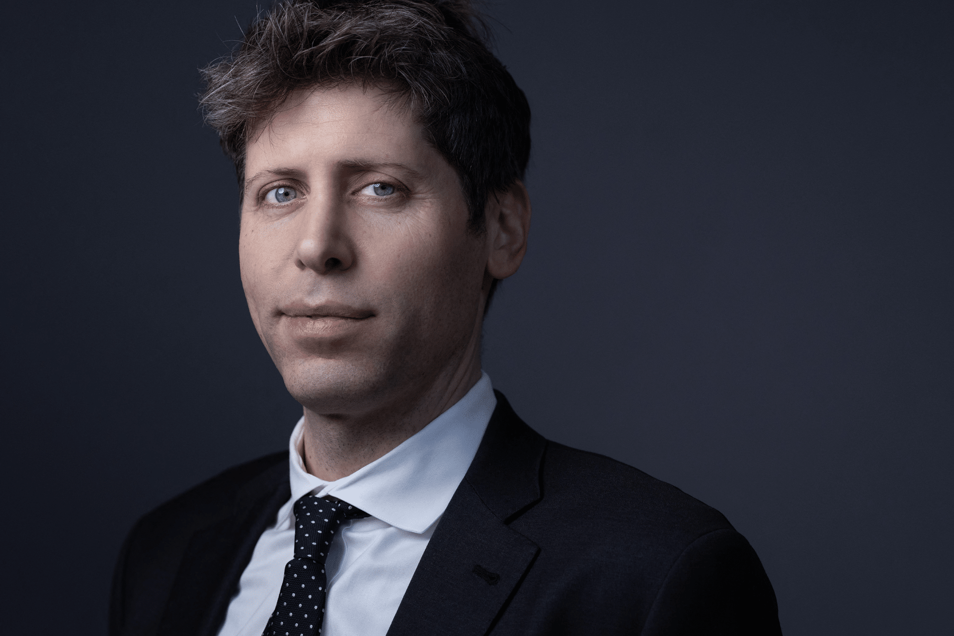 A man with light skin, short brown hair with some gray, and blue eyes—resembling Sam Altman of rocket company fame—wears a black suit, white shirt, and black polka dot tie while posing against a dark gray background.