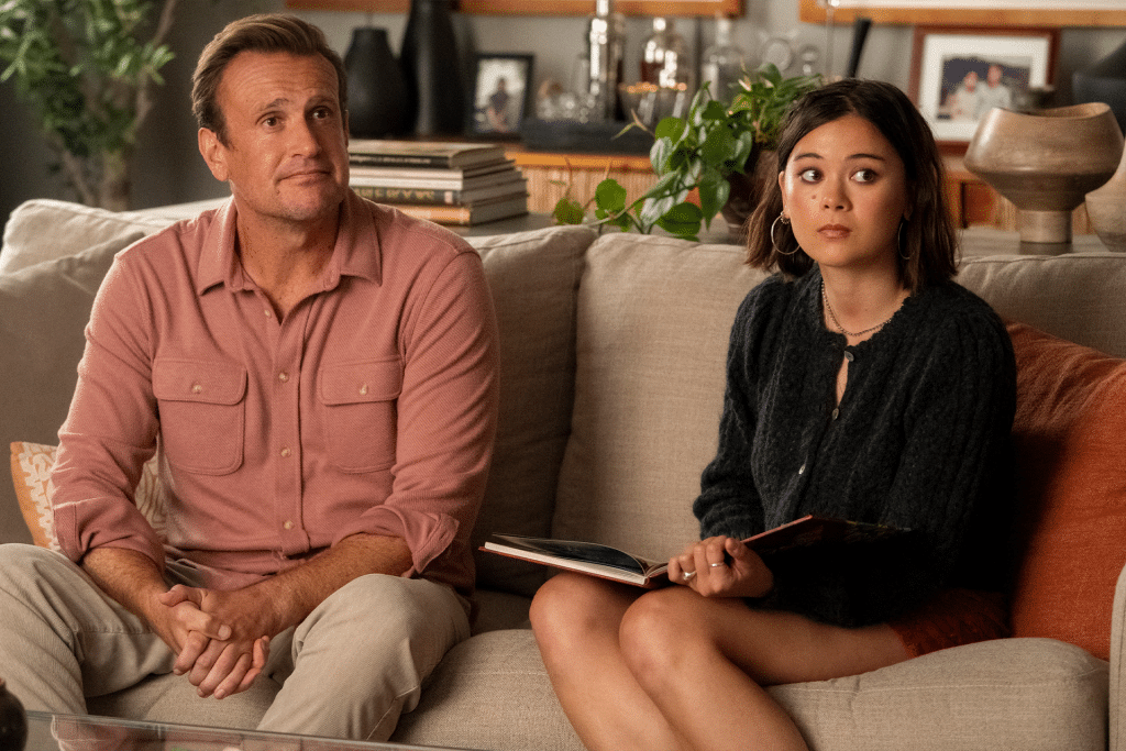 A man in a pink shirt and a young woman holding a book sit together on a beige couch, looking slightly off to the side—perhaps discussing Shrinking season 3 in their cozy living room filled with plants and decor.