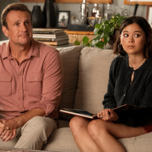 A man in a pink shirt and a young woman holding a book sit together on a beige couch, looking slightly off to the side—perhaps discussing Shrinking season 3 in their cozy living room filled with plants and decor.