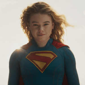 A woman with blonde wavy hair stands confidently, wearing a blue superhero suit with a red and yellow "S" emblem and red cape. Sunlight shines behind her against a cloudy sky, capturing the essence of the Supergirl trailer first look.