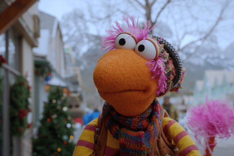 A colorful puppet with orange skin, wide eyes, and pink fuzzy hair wears a knitted hat and striped scarf, standing outdoors in a wintery town scene inspired by a Fraggle Rock holiday special.