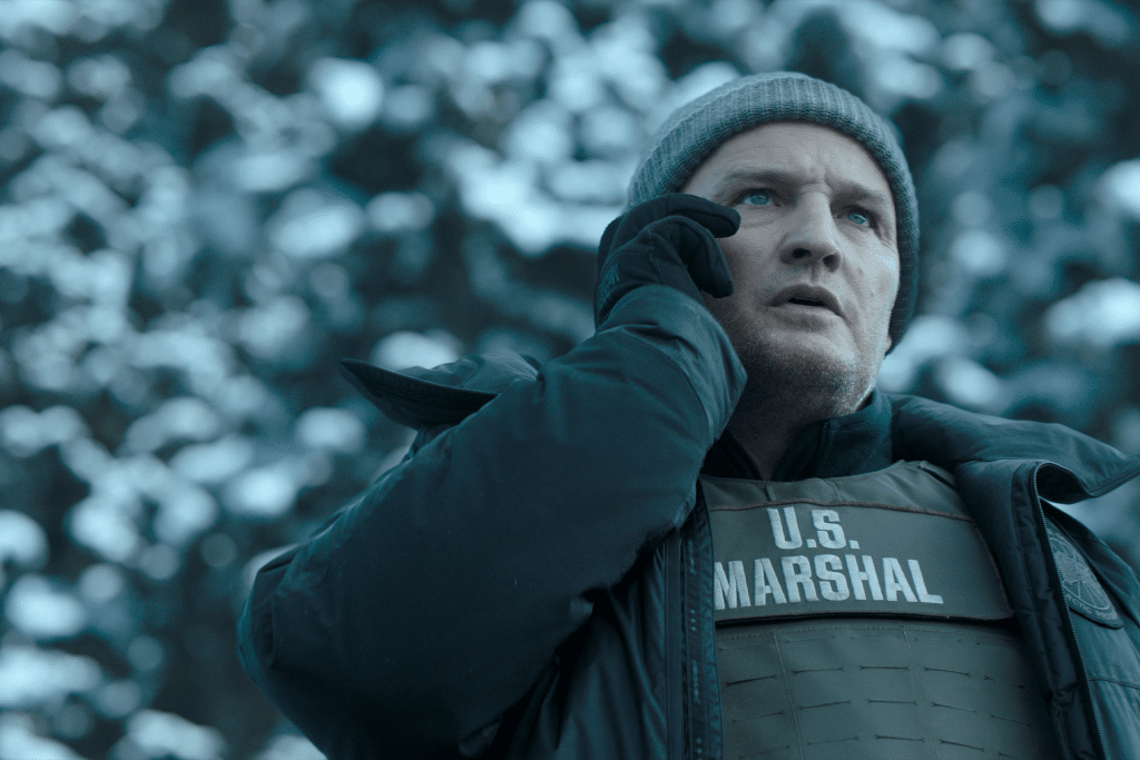 A man in a winter beanie and jacket, wearing a bulletproof vest labeled "U.S. Marshal," stands outdoors in a snowy environment, holding a phone to his ear and looking concerned—perhaps awaiting news on Apple TV’s decision to cancel The Last Frontier.