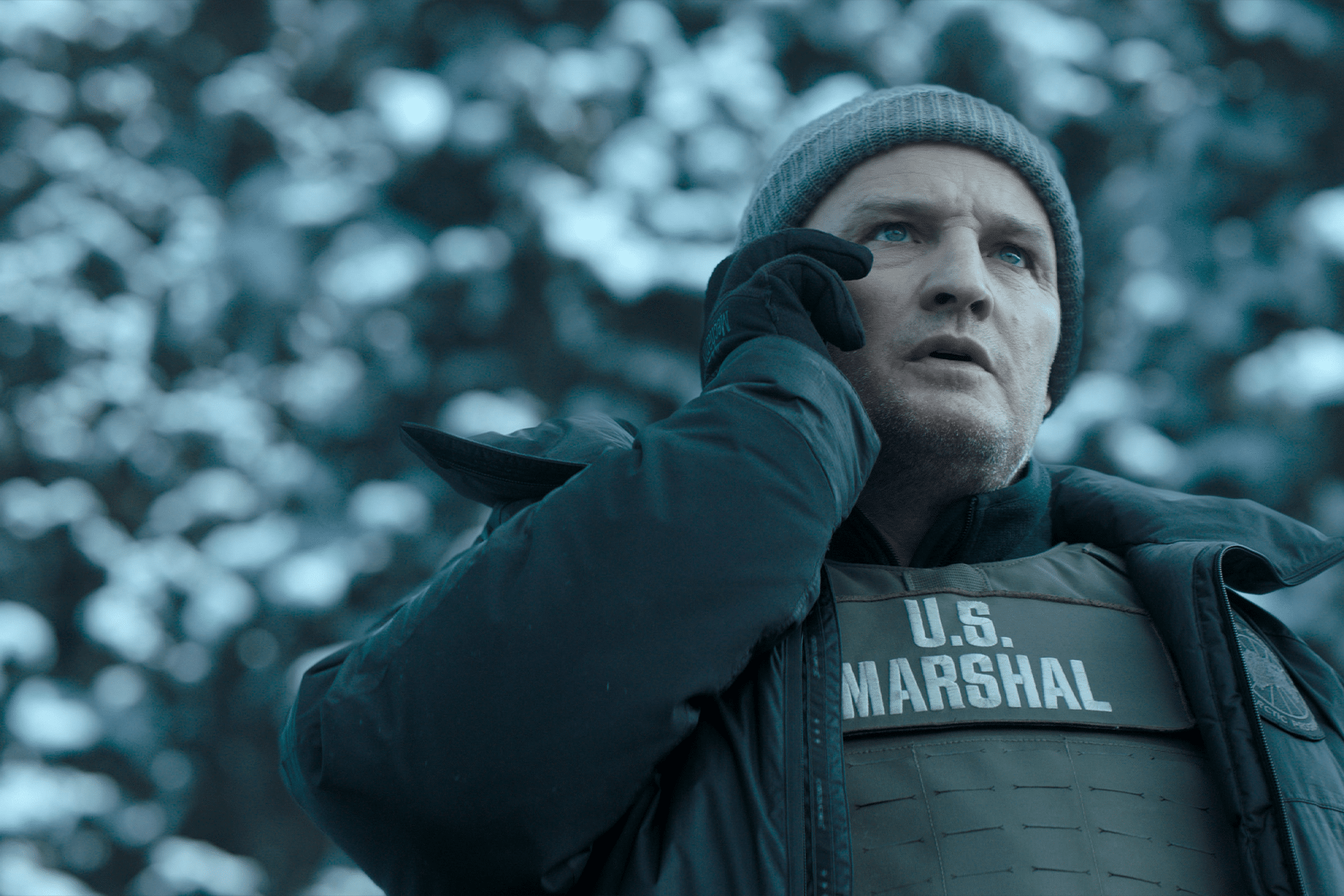 A man in a winter beanie and jacket, wearing a bulletproof vest labeled "U.S. Marshal," stands outdoors in a snowy environment, holding a phone to his ear and looking concerned—perhaps awaiting news on Apple TV’s decision to cancel The Last Frontier.