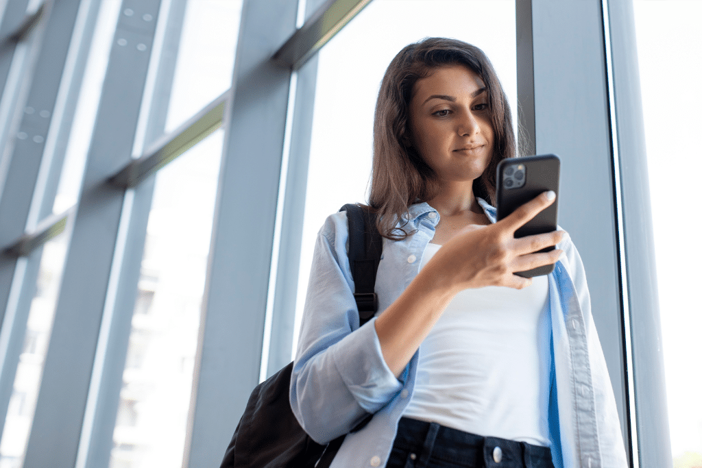A young woman with long dark hair, wearing a light blue shirt and backpack, stands by large windows and looks at her iPhone with a slight smile, perhaps reviewing travel safety tips for her next adventure.