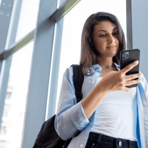 A young woman with long dark hair, wearing a light blue shirt and backpack, stands by large windows and looks at her iPhone with a slight smile, perhaps reviewing travel safety tips for her next adventure.