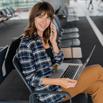 Apple Travel - A woman sits in an airport terminal, smiling while talking on her iPhone. She has a laptop on her lap, wears a plaid shirt and brown pants, and there are empty seats around her as she checks travel safety tips.