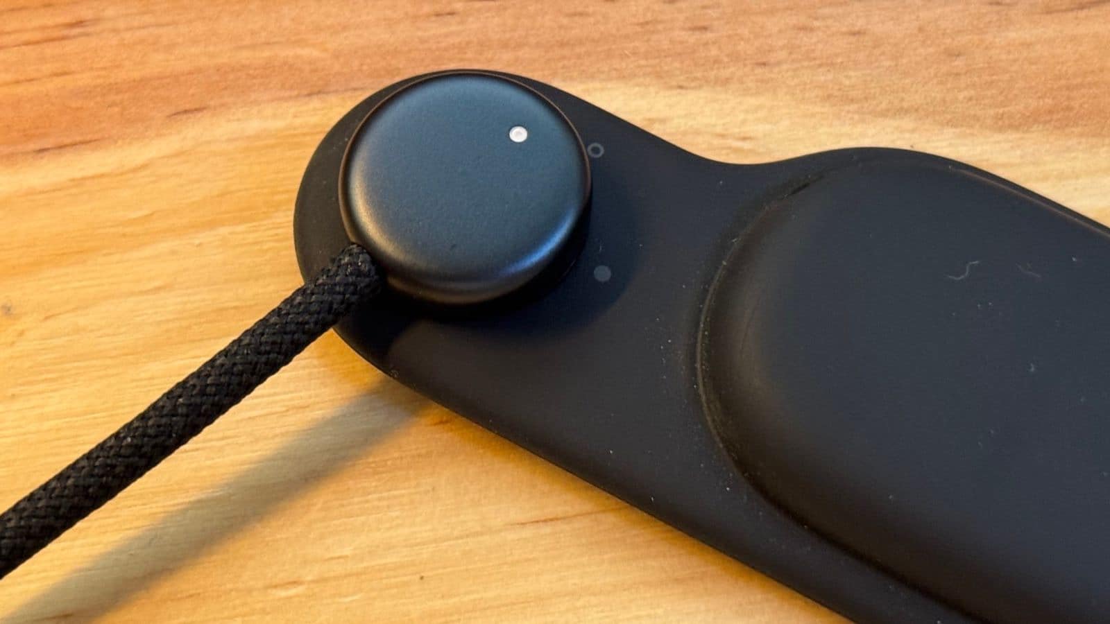 Close-up of an all-black charging puck with a fabric cable attached, resting on a matching black charging pad atop a wooden surface—reminiscent of Vision Pro prototype parts.