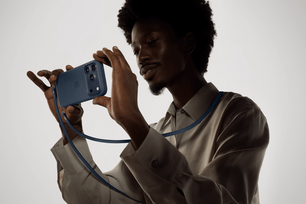 A person wearing a light-colored shirt holds a blue smartphone with an apple phone case horizontally, both hands gripping it. The matching blue strap hangs around their neck against a plain, light-colored background.