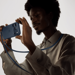 A person wearing a light-colored shirt holds a blue smartphone with an apple phone case horizontally, both hands gripping it. The matching blue strap hangs around their neck against a plain, light-colored background.