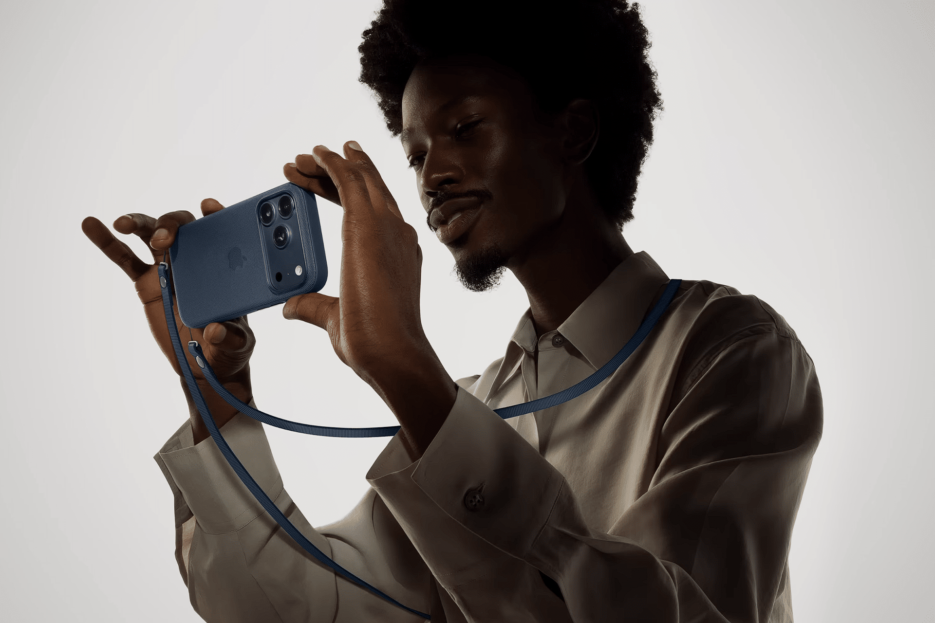 A person wearing a light-colored shirt holds a blue smartphone with an apple phone case horizontally, both hands gripping it. The matching blue strap hangs around their neck against a plain, light-colored background.