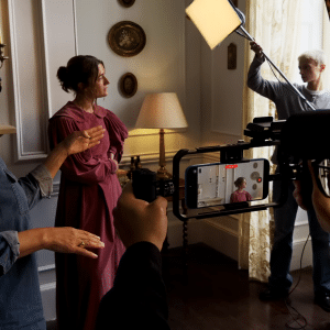 Two people converse on a film set, one in a pink costume. Crew members use lighting equipment, sound boom, and an iPhone 17 Pro for video making. The indoor scene features candles, framed pictures, and a lamp in the background.