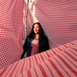 A woman stands among billowing red and white striped and checkered fabric panels outdoors, with sunlight casting shadows—perfect inspiration for holiday family photography. She wears a denim jacket over a pink striped shirt and smiles softly.