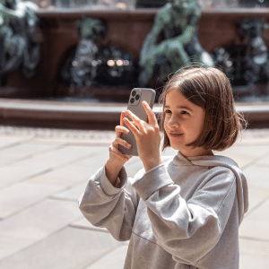 A young girl in a light gray hoodie stands outdoors near a fountain, smiling as she takes a photo or video with her iPhone for kids safety. Statues and flowing water are visible in the blurred background.