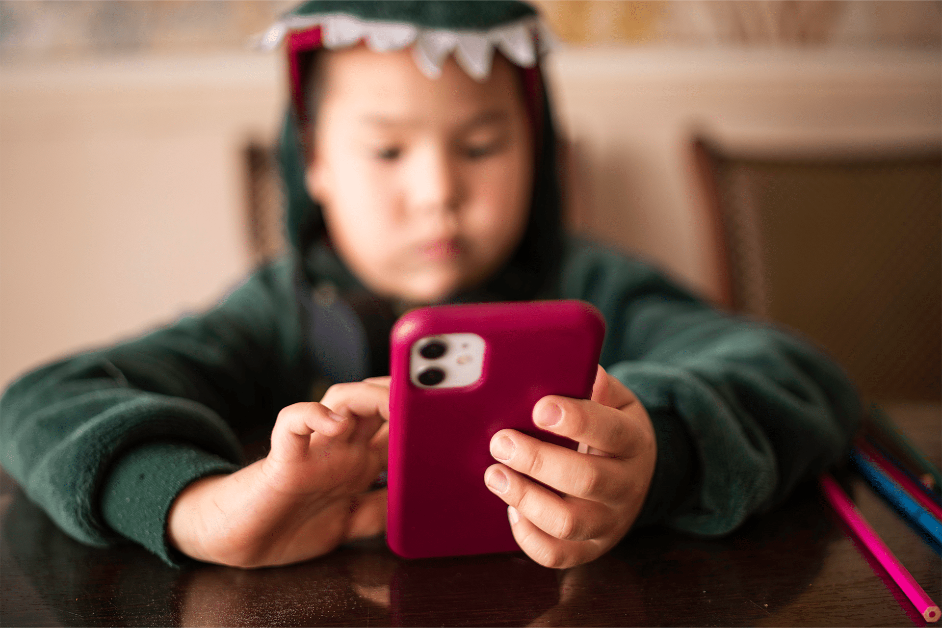 Parent Digital Routine - A child wearing a green dinosaur hoodie sits at a table, using a pink smartphone with both hands. The focus is on the phone and hands, highlighting iphone for kids safety, while the child’s face is blurred in the background.