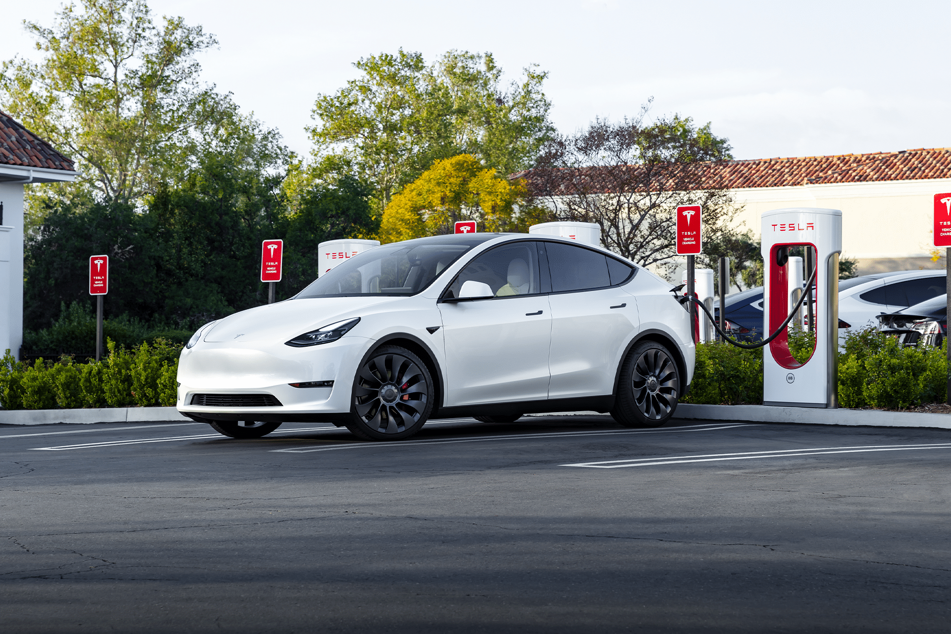 A white Tesla electric car is parked at a Tesla Supercharger station at a new location in Florida, plugged in and charging, with other charging stations and greenery in the background.