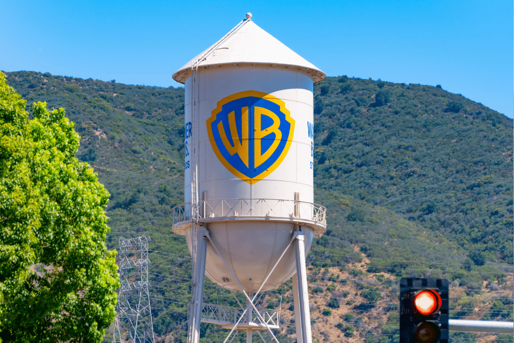 A large white water tower with the blue and yellow Warner Bros. logo stands in front of green hills under a clear blue sky, symbolizing the studio’s legacy amid recent headlines like the Netflix Warner Deal. A leafy tree and red traffic light are in the foreground.