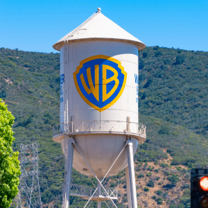 A large white water tower with the blue and yellow Warner Bros. logo stands in front of green hills under a clear blue sky, symbolizing the studio’s legacy amid recent headlines like the Netflix Warner Deal. A leafy tree and red traffic light are in the foreground.