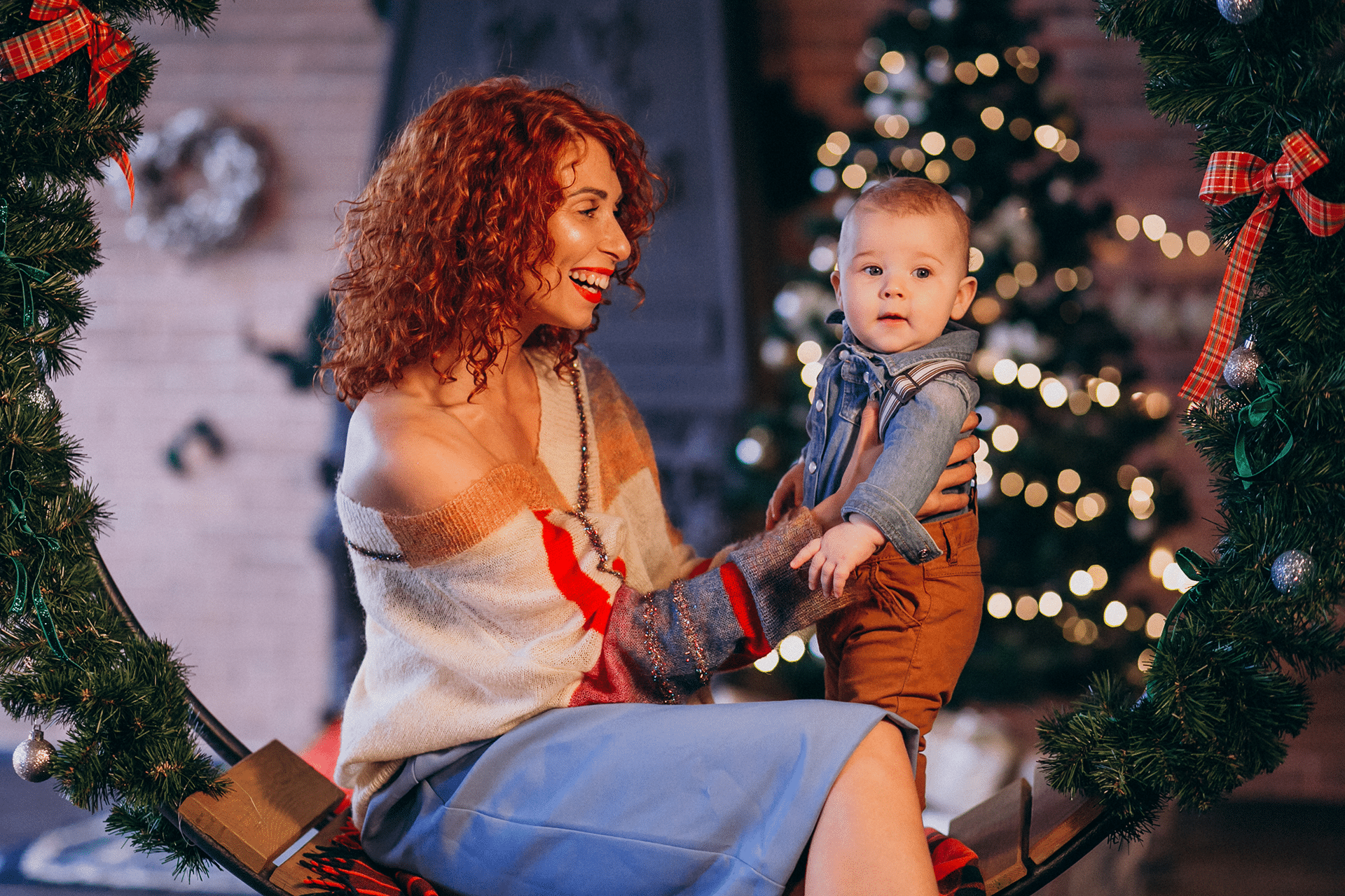 A smiling woman with curly red hair sits on a swing decorated with greenery and bows, holding a baby in suspenders. Holiday family photography captures the warmth of festive lights and a Christmas tree using cozy indoor lighting.