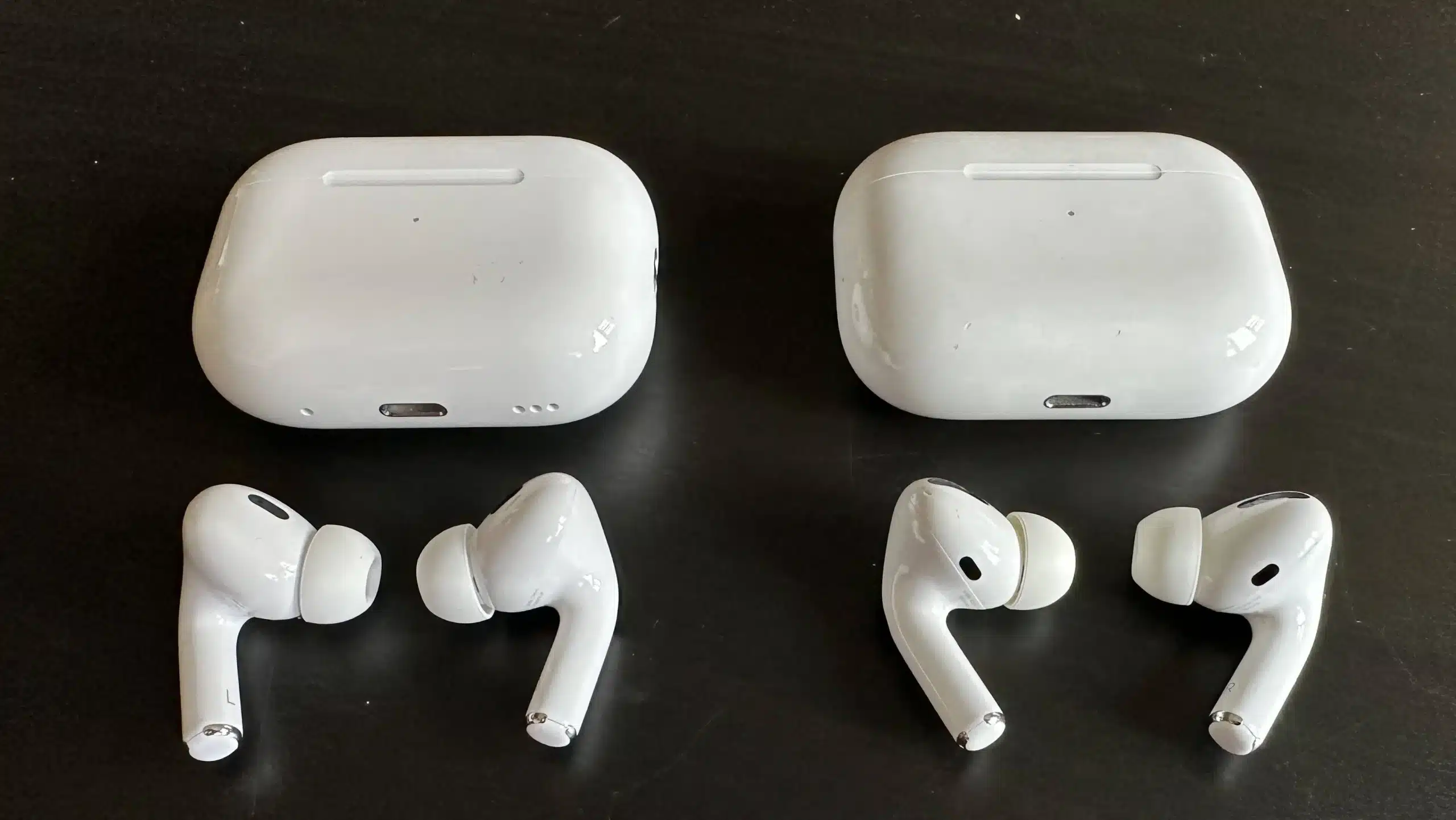 Two pairs of white wireless earbuds, resembling Counterfeit AirPods Pro, are placed with their charging cases on a black surface. Each pair is positioned in front of its respective case, with the earbuds facing outward.